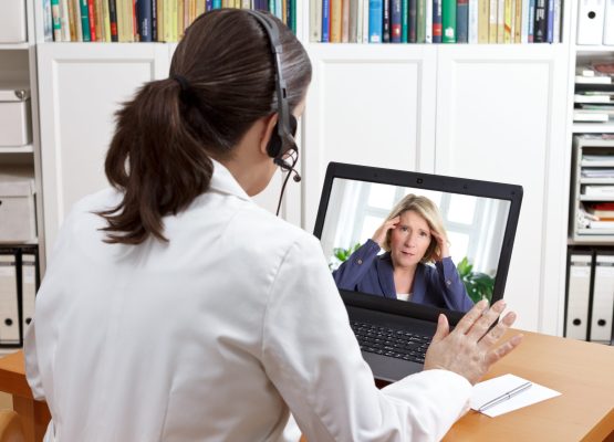 Female doctor or pharmacist in her surgery office with headset in front of her computer talking via video chat with a patient, telling her to take things easy